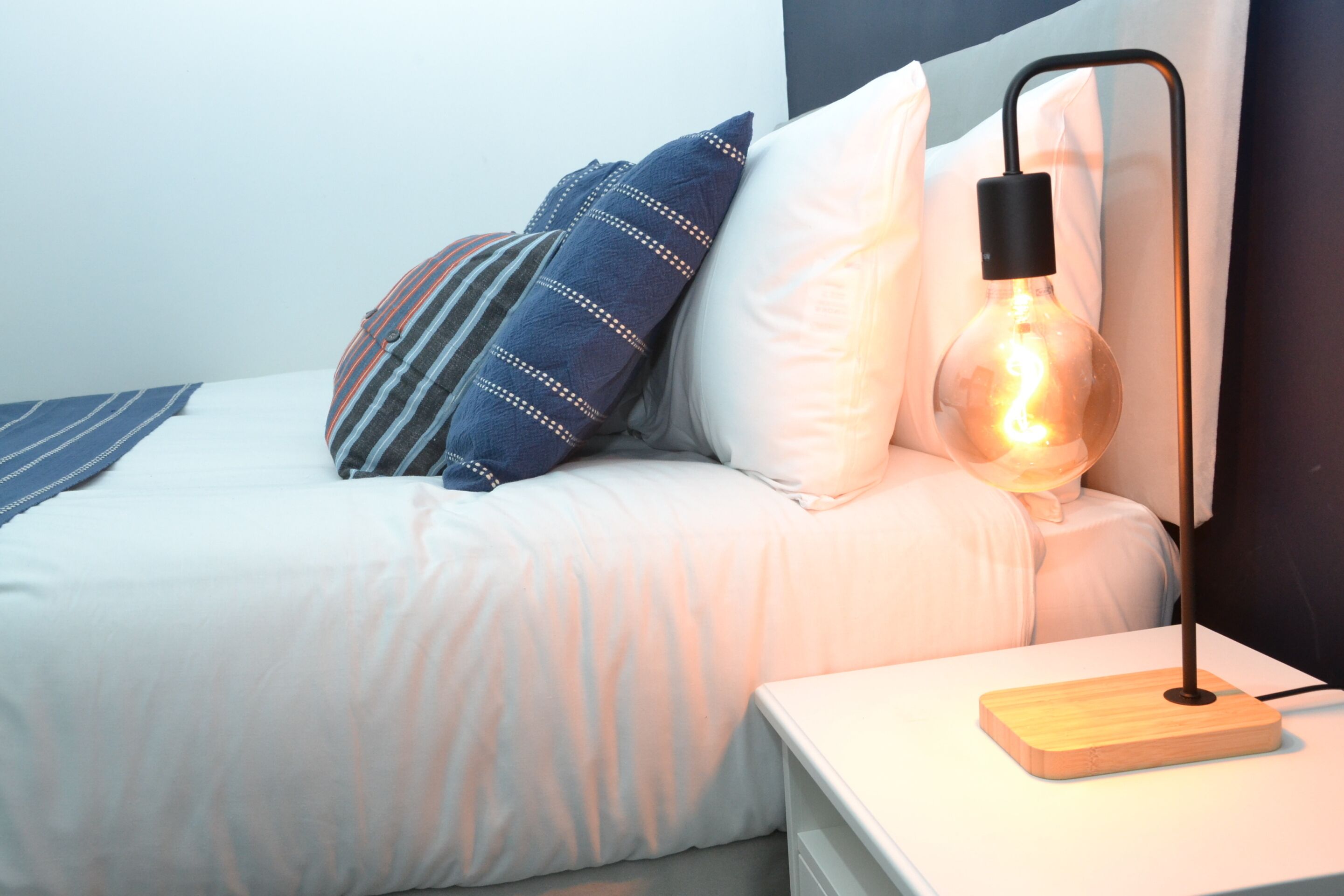 A neatly made bed with white bedding, a striped pillow, and a light bulb lamp on a bedside table.