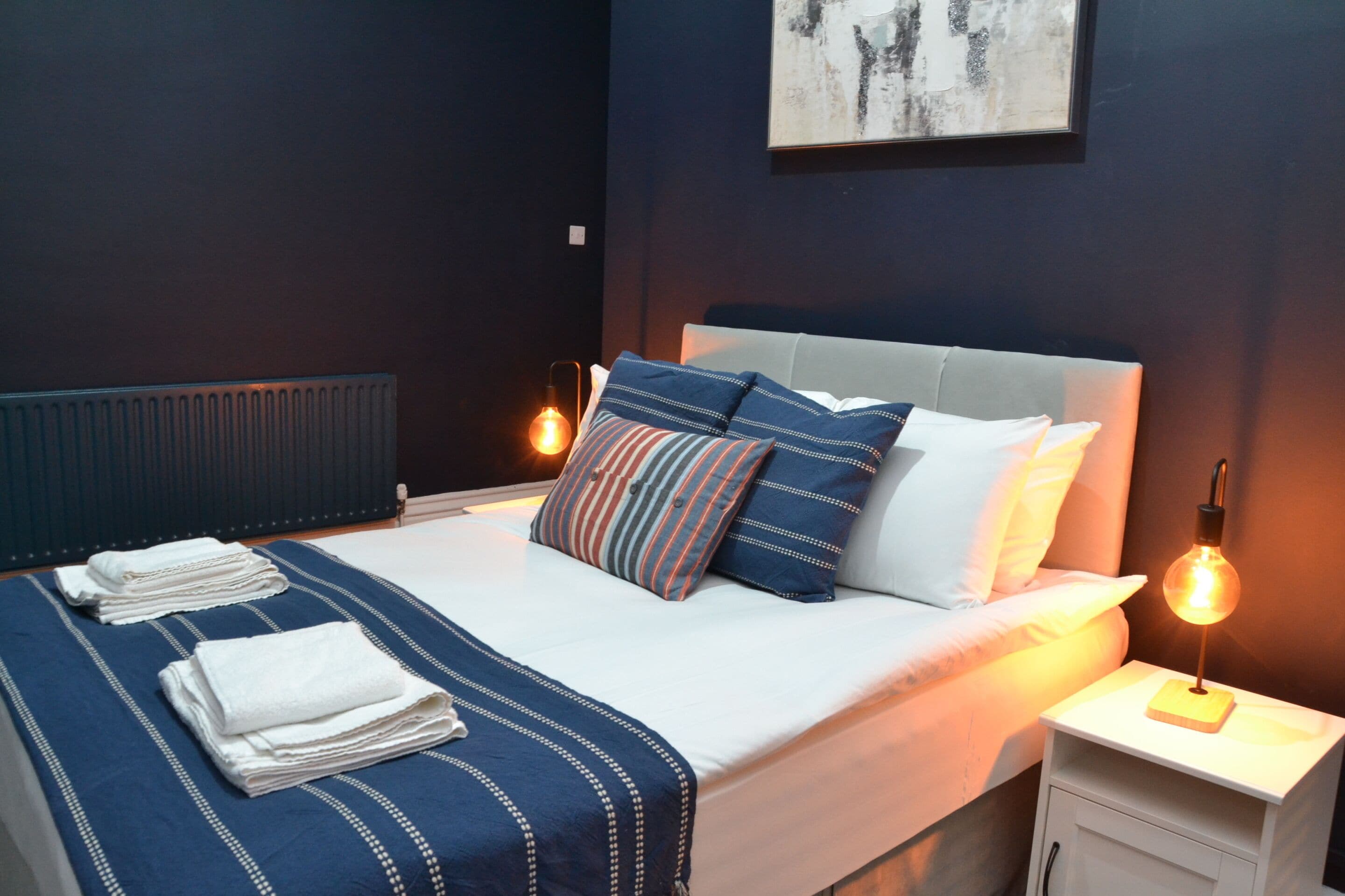 A neatly made bed with a blue and white bedspread, a striped pillow, and a white pillow. There is a small bedside table with a lamp.