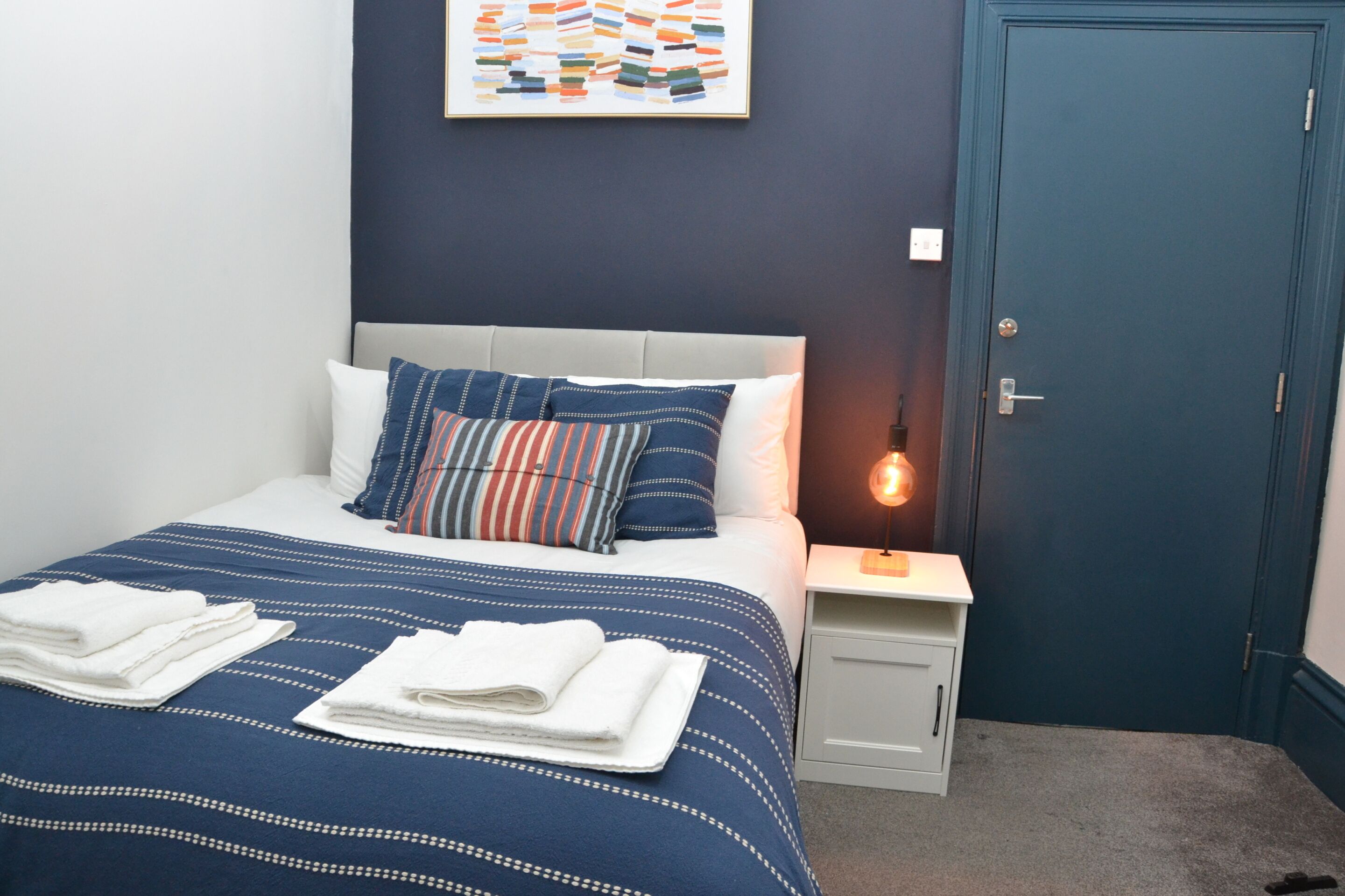 A neatly made bed with a striped bedspread and pillows, a bedside table with a lamp, and a door to the right.