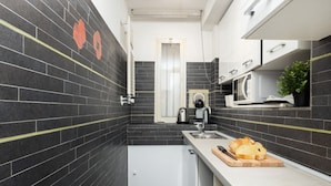 A modern kitchen with black tiled walls, a white countertop, a sink, a kettle, and a loaf of bread on a cutting board. - Cozy 45m2 apartment, ideal for 4 people, located on the ground floor of a building.The accommodation is located in a residential area 2 steps from the famous Prada Foundation, center of culture and fashion, and a short distance from the Milano Porta Rom (Milan)