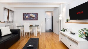 A modern living room with a black sofa, white dining table, and chairs, a TV mounted on the wall, and a painting above the table. - Cozy 45m2 apartment, ideal for 4 people, located on the ground floor of a building.The accommodation is located in a residential area 2 steps from the famous Prada Foundation, center of culture and fashion, and a short distance from the Milano Porta Rom (Milan)
