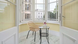 Een kamer met erker, een rieten stoel, een tafeltje met een vaas en een kopje, en een witte deur.