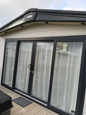 A building with a large, black-framed door with a window, a sign with 'ABI' logo, and a small outdoor area with a table and a mat. - Holiday Park Home With Private Hot Tub (Cirencester)