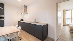 Eine moderne Küche mit einer zentralen Kochinsel, einem Esstisch mit Stühlen und Blick in ein Schlafzimmer mit Bett und Vorhängen.