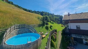 Une piscine entourée d’une clôture en bois, une maison avec un toit en tuiles et, au loin, un paysage vallonné.