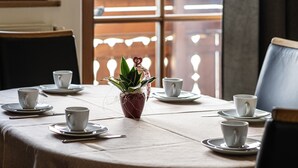 Une table de salle à manger ronde, avec des tasses et des soucoupes blanches, une plante en pot et une vue par une fenêtre ornée d’un treillis en bois.