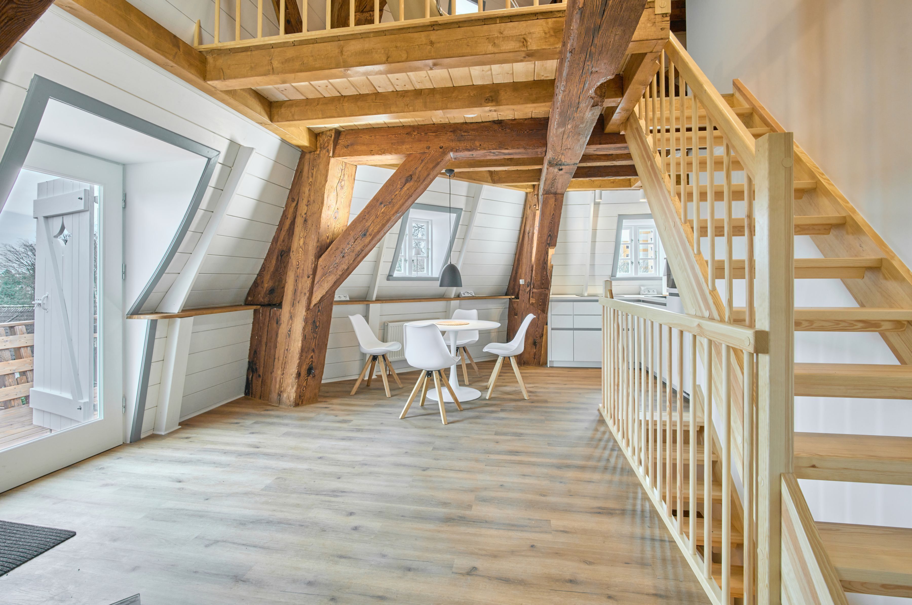 Una habitación estilo loft con vigas de madera a la vista, un comedor con sillas blancas y una escalera de madera.