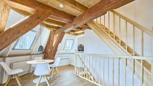 A loft-style room with exposed wooden beams, a dining area with white chairs, and a kitchen in the background. - fewo1846 - Westwind - Vacation in a historic windmill with max. 4 persons (Flensburg)