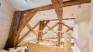 A room with exposed wooden beams, a bed with a geometric-patterned bedspread, and a view of the upper floor through a wooden railing. - fewo1846 - Westwind - Vacation in a historic windmill with max. 4 persons (Flensburg)