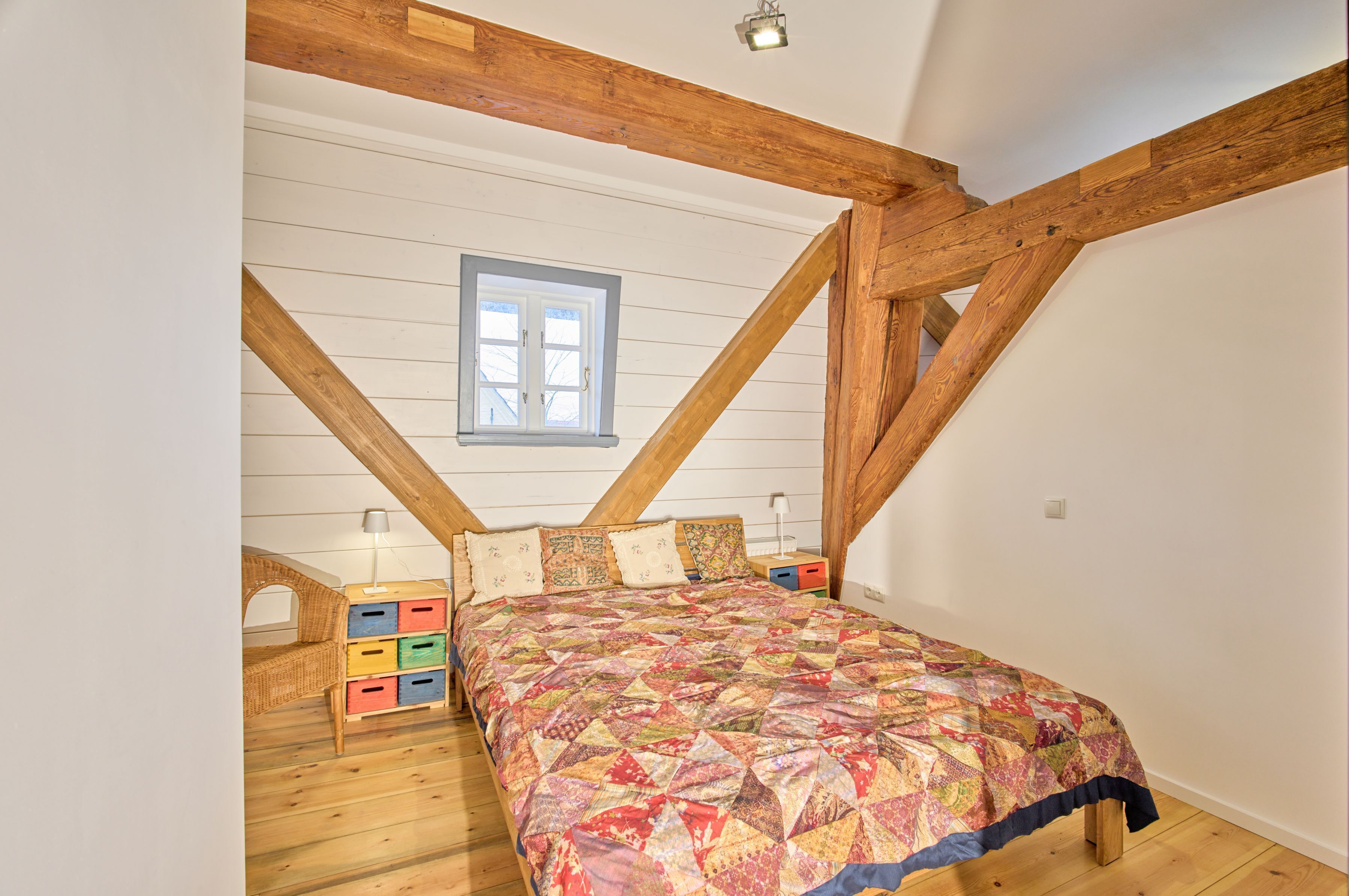 A bedroom with a wooden ceiling, a window, a bed with a colorful quilt, a nightstand, and a dresser with drawers.