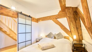 A bedroom with a wooden ceiling, a bed with a white comforter, two bedside tables with lamps, and a sliding glass door. - Apartment 1846 - Ostwind - Vacation in a historic windmill for up to 4 people (Flensburg)