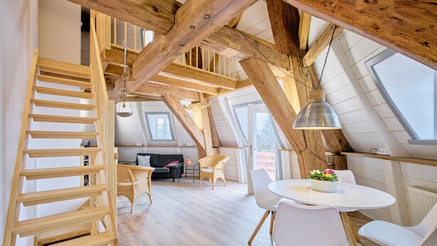A loft-style room with exposed wooden beams, a wooden ladder, white furniture, and large windows. - Apartment 1846 - Ostwind - Vacation in a historic windmill for up to 4 people (Flensburg)