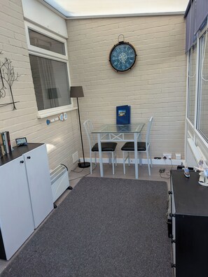 A room with a brick wall, a round wall clock, a table with chairs, a small cabinet, and a radiator. - Bungalow Sleeps 4 With Parking (Skegness)