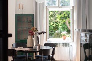 Ein Essbereich mit Tisch, Stühlen, einer Blumenvase, einem grünen Schrank und einem Fenster mit Blick ins Grüne.