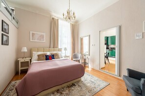 A bedroom with a bed, bedside tables, a chair, a rug, a chandelier, and a view of the kitchen through an open door. - Riverside 2BR Apartment Next to the National Theatre by EasyBNB Premium (Prague)