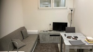 Un salon moderne avec un canapé gris, un téléviseur, une petite table avec une lampe et une fenêtre au-dessus.