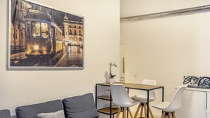 Ein modernes Wohnzimmer mit einem grauen Sofa, einem Esstisch mit Stühlen, einer kleinen Pflanze und einem gerahmten Bild einer Straßenbahn.