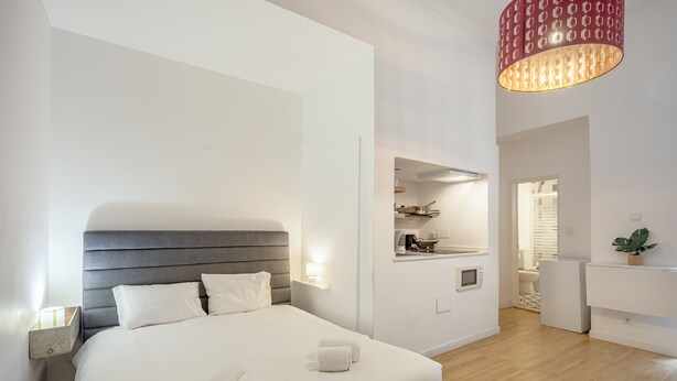 Ein modernes Hotelzimmer mit einem großen Bett, Nachttischen, einem kleinen Schreibtisch, einem Spiegel und einer roten, gemusterten Leuchte.