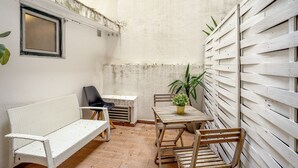 Eine kleine Terrasse mit einer weißen Rattan-Couch, einem Holztisch mit Stühlen, einem Blumentopf und einem Fenster.