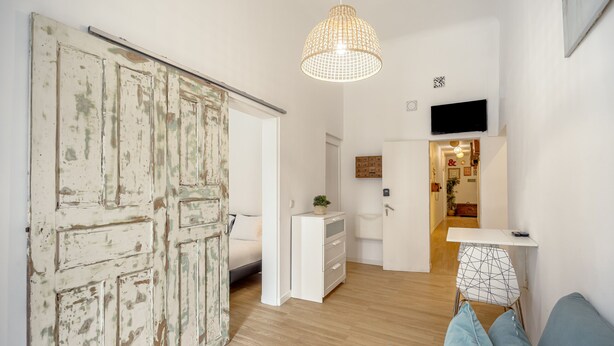 Um quarto com porta deslizante, uma cama, escrivaninha com gavetas, luminária pendente e televisão na parede.