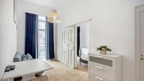 Quarto moderno com escrivaninha branca, cómoda branca, luminária pendente e vista através de uma porta.