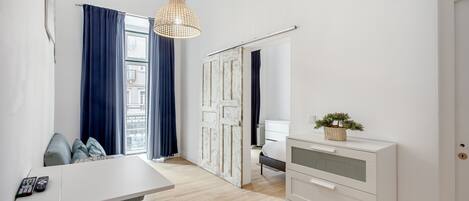 A modern room with a white desk, a white dresser, a hanging light fixture, and a view through a door.