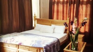 A wooden bed with a headboard, a bedside table with a vase of flowers, and a window with curtains.