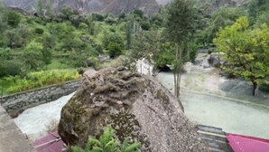 A mountainous landscape with a large rock formation in the foreground, a stone wall, and a small building with a red roof.