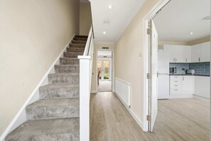 Eine Treppe führt zu einer Tür, eine Küche mit weißen Schränken und einer blauen Spritzschutzwand, und ein Flur mit hellem Holzboden.