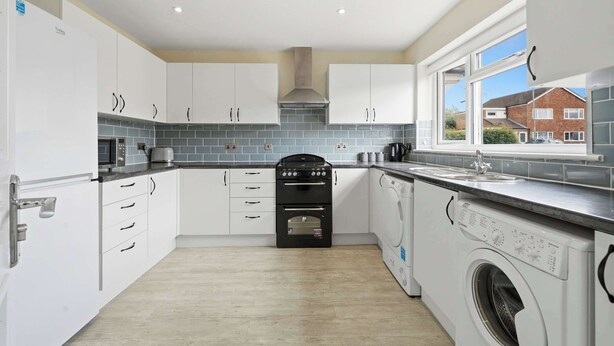 Une cuisine moderne avec des meubles blancs, un four noir, un lave-linge et une fenêtre donnant sur un immeuble en briques.