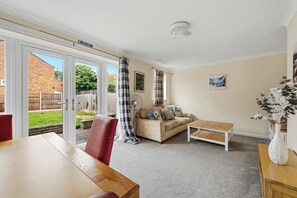 Ein Wohnzimmer mit einer Couch, einem Holztisch, einer weißen Vase mit Blumen, einem Ventilator und Blick ins Freie.