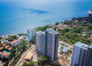 Un immeuble de grande hauteur, doté de nombreux étages, entouré de verdure et situé près d’une zone côtière avec une plage et une étendue d’eau.