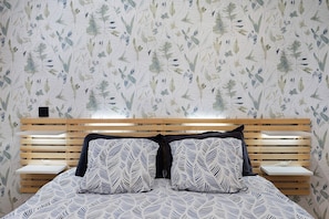 Un dormitorio con cama, cabecero de madera, mesitas de noche y papel tapiz estampado.