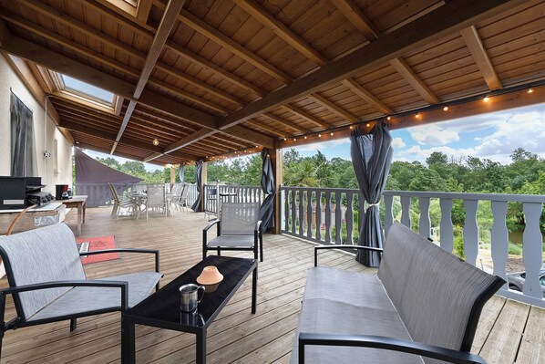 A covered outdoor deck with wooden flooring, seating arrangements, and a view of the surrounding greenery.