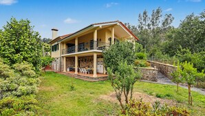 Una casa de dos pisos con balcón, rodeada de vegetación y un muro de piedra.