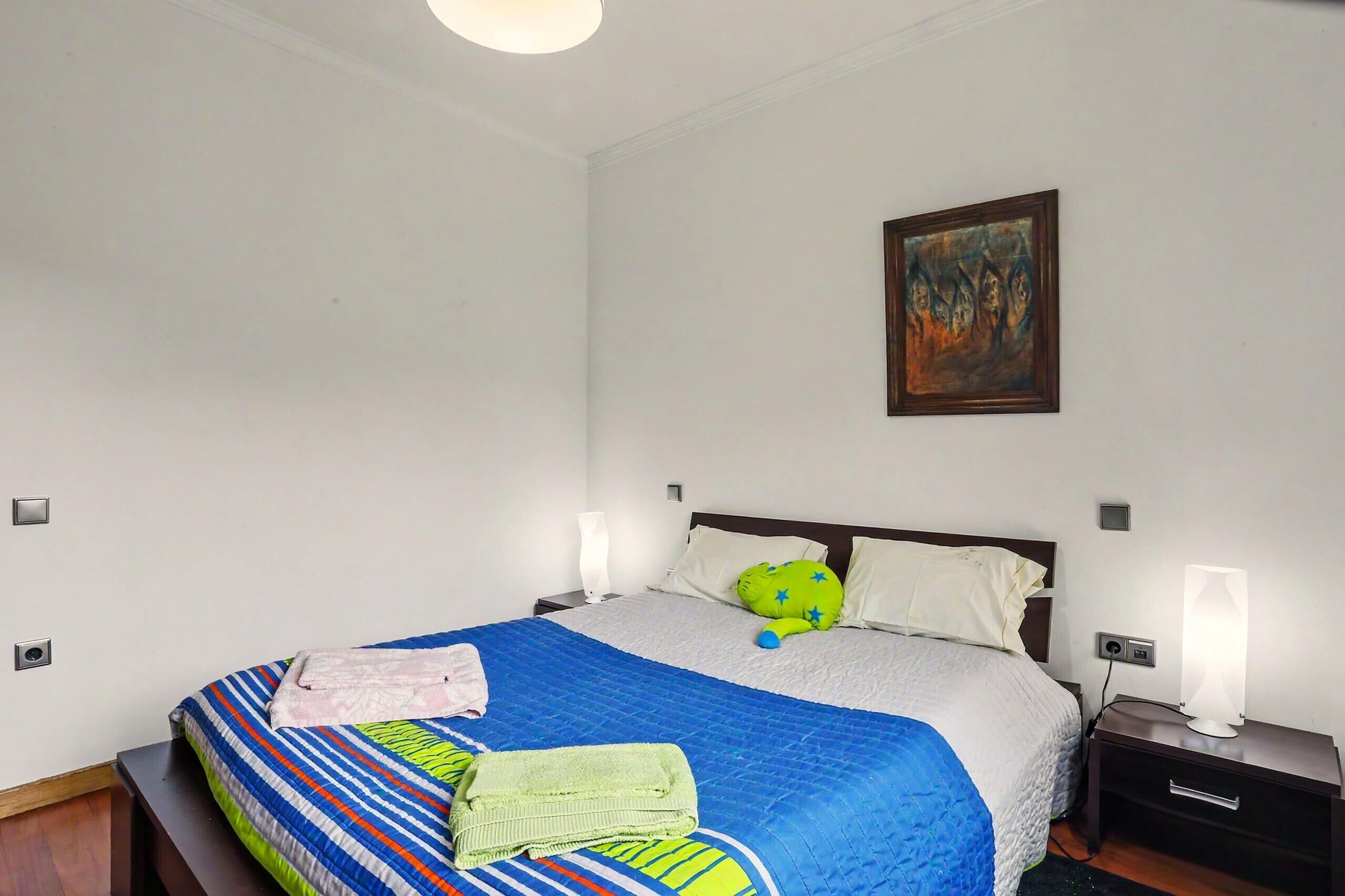 A neatly made bed with a striped bedspread, a green towel, and a blue stuffed toy. There is a painting on the wall and a bedside table with a lamp.