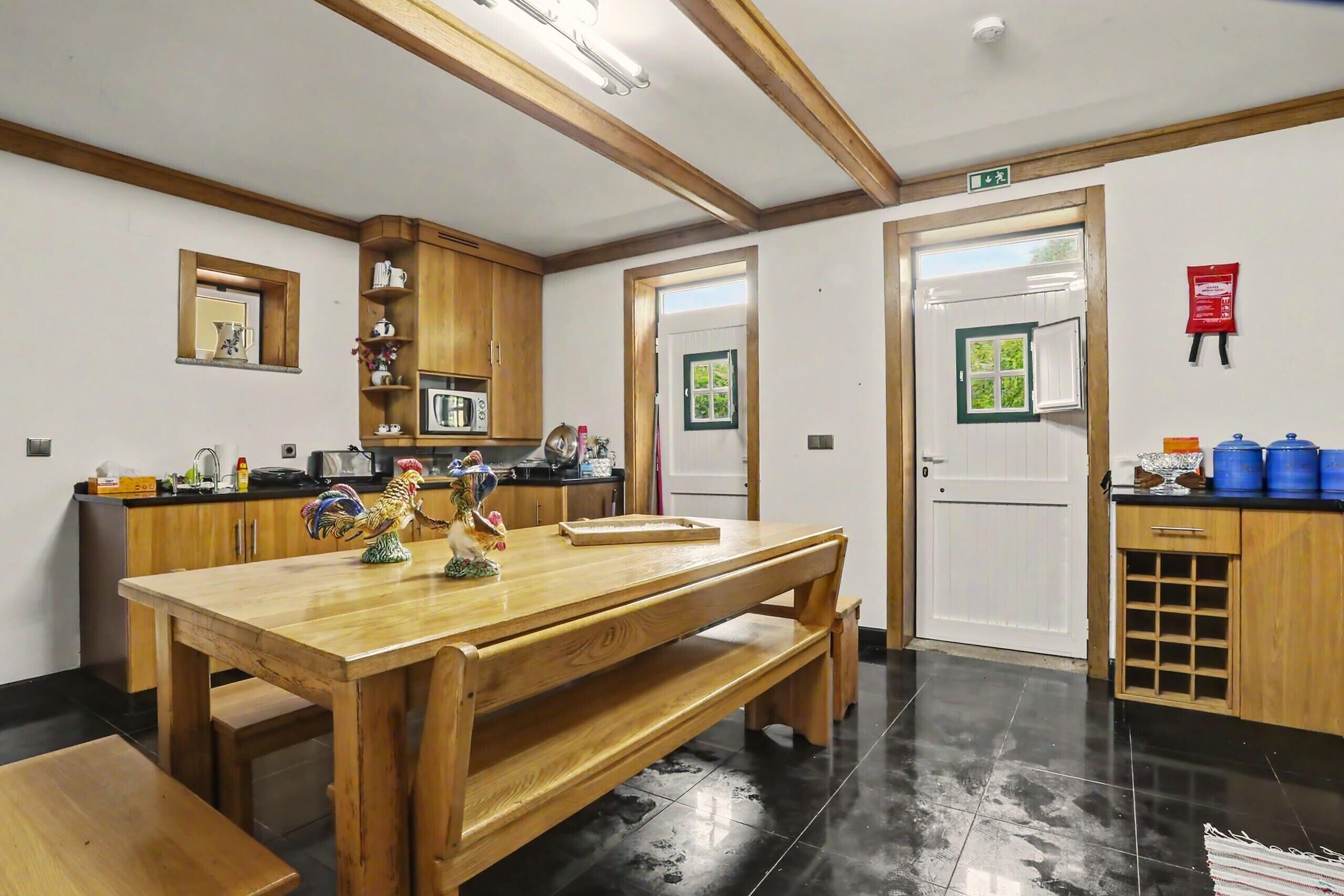Cozinha com armários de madeira, uma mesa de madeira e um banco. Há uma porta com uma janela verde, um extintor de incêndio e uma pequena prateleira com objetos.