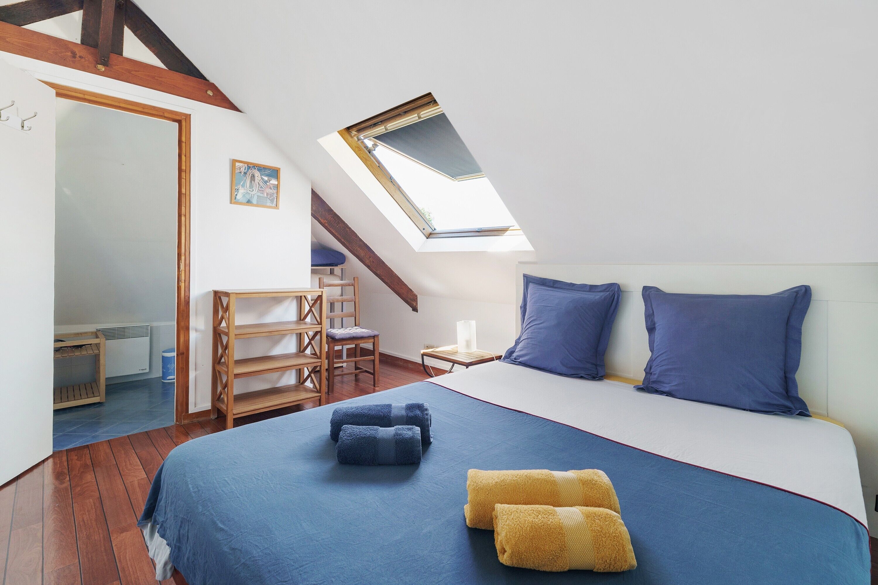 Une chambre avec un puits de lumière, un lit avec une literie bleue et blanche, une chaise en bois et une petite table.