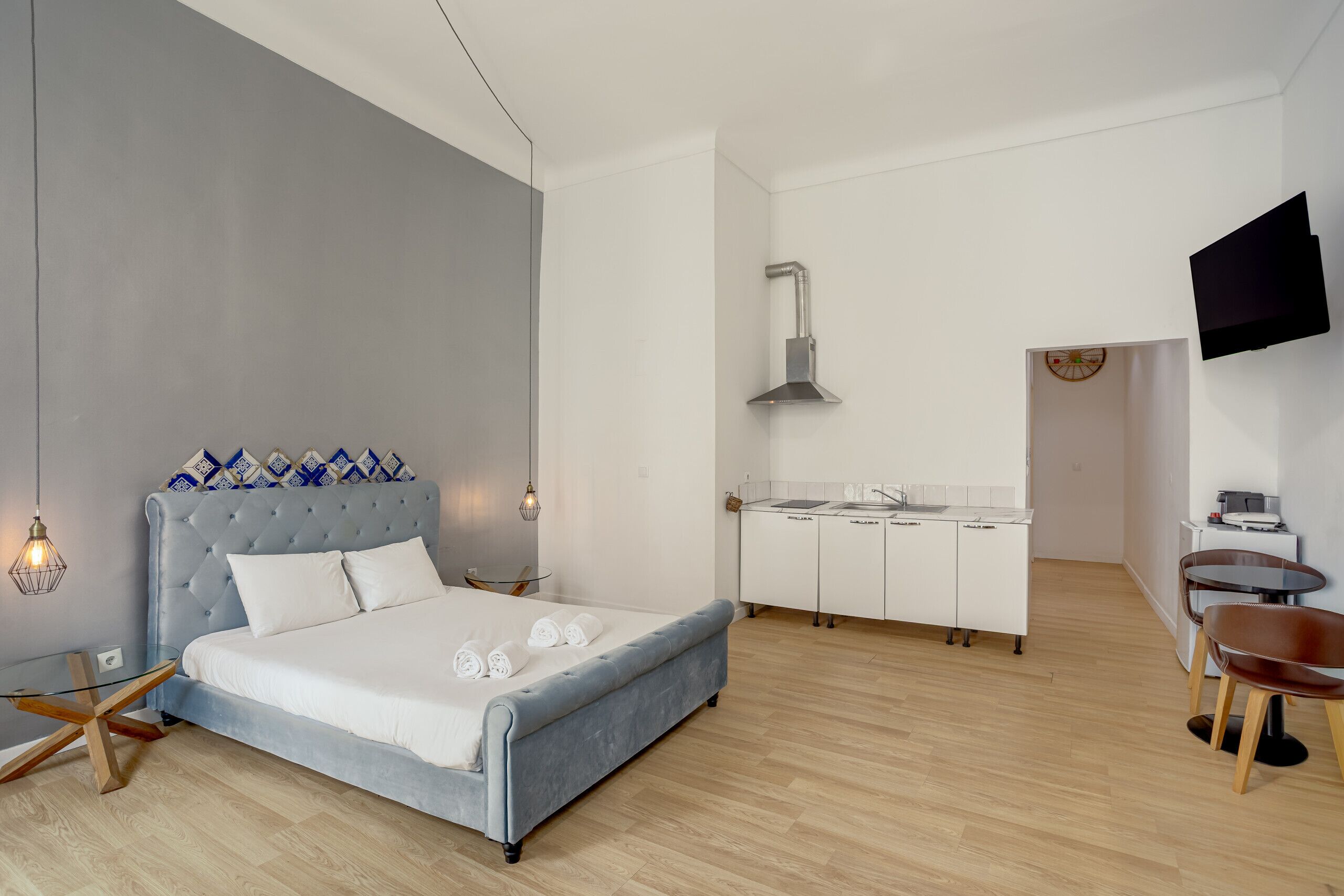 A modern bedroom with a large bed, a small kitchenette, a TV, and a wooden floor.