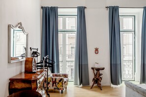 Ein Zimmer mit Schreibtisch aus Holz, einem Stuhl, einer gemusterten Couch, einem kleinen Tisch und einem großen Fenster mit Vorhängen.