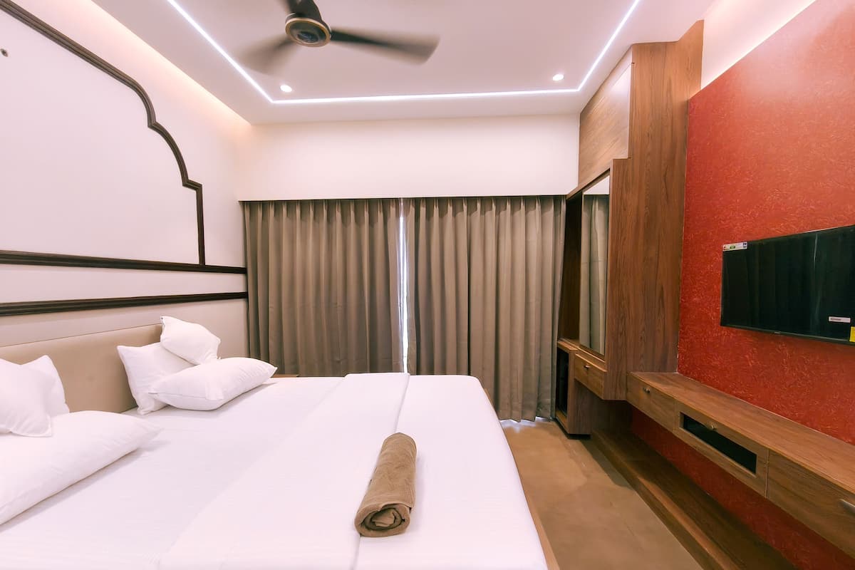 Una habitación de hotel con cama, ventilador de techo, televisor montado en la pared y un gran ventanal con cortinas.