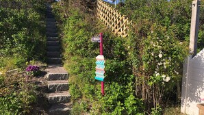 Uma escadaria que sobe, cercada por vegetação exuberante e uma placa indicando a direção da praia.
