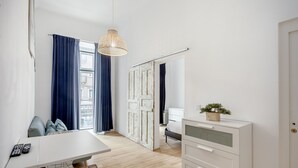 Habitación de hotel moderna con zona de comedor, cómoda blanca, planta pequeña y vista a través de una puerta.