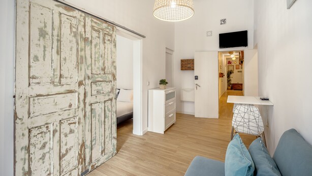 Una habitación con puerta corrediza, una cama, un aparador, una mesita con una silla de estampado geométrico y una lámpara colgante.