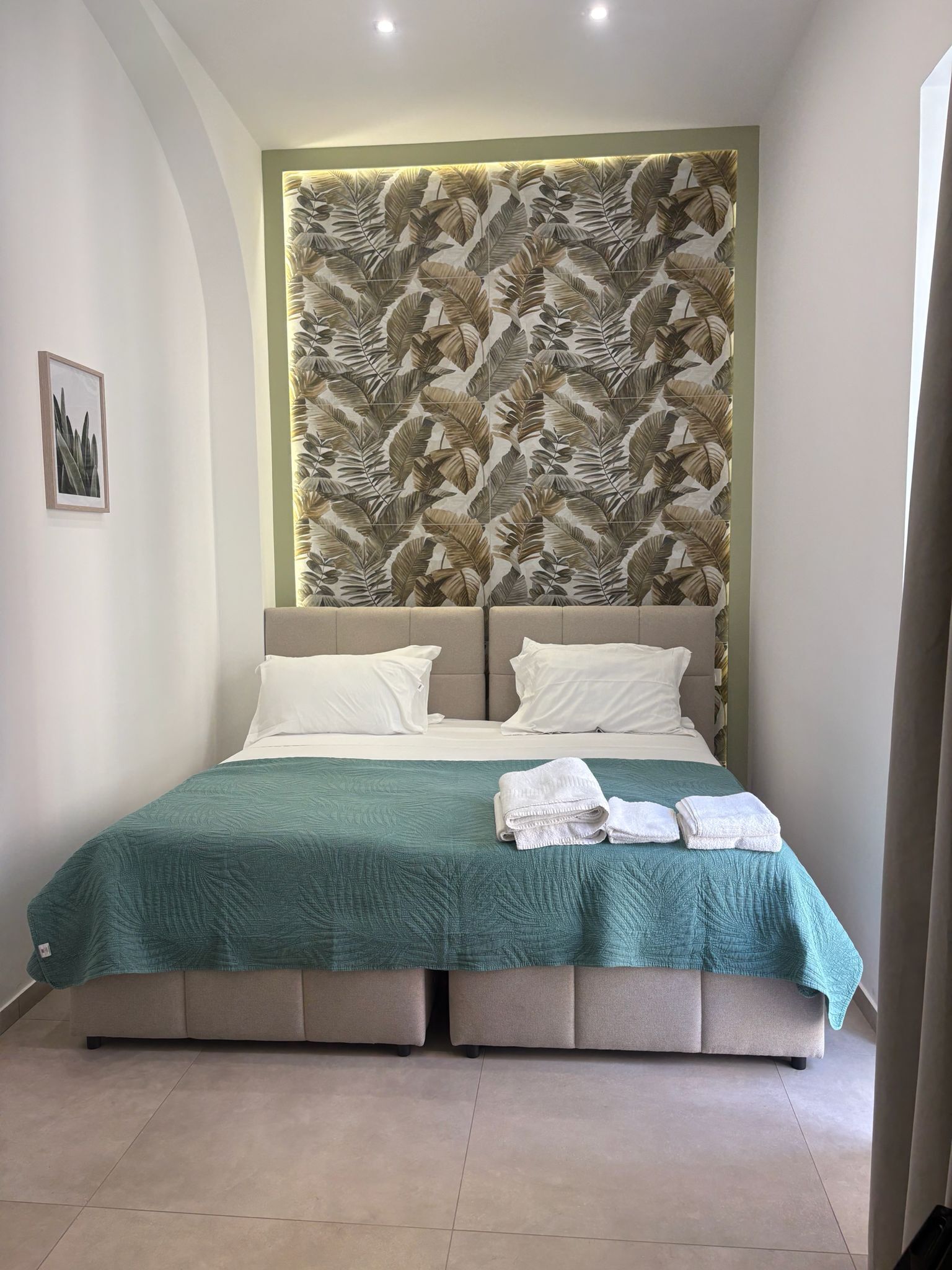 Una cama doble con cubrecama verde, dos almohadas blancas y un cabecero con estampado de hojas.