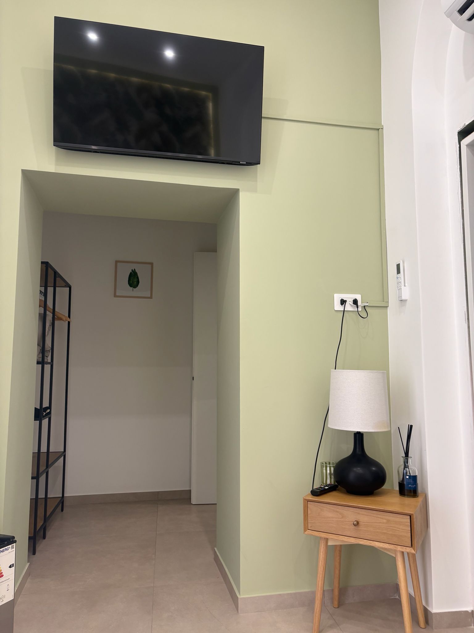 A room with a flat-screen TV mounted on the wall, a wooden bedside table with a lamp, and a power outlet on the wall.