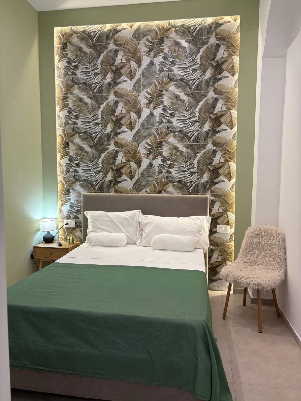 Un dormitorio con cama, mesita de noche, lámpara, silla y pared con diseño de hojas.