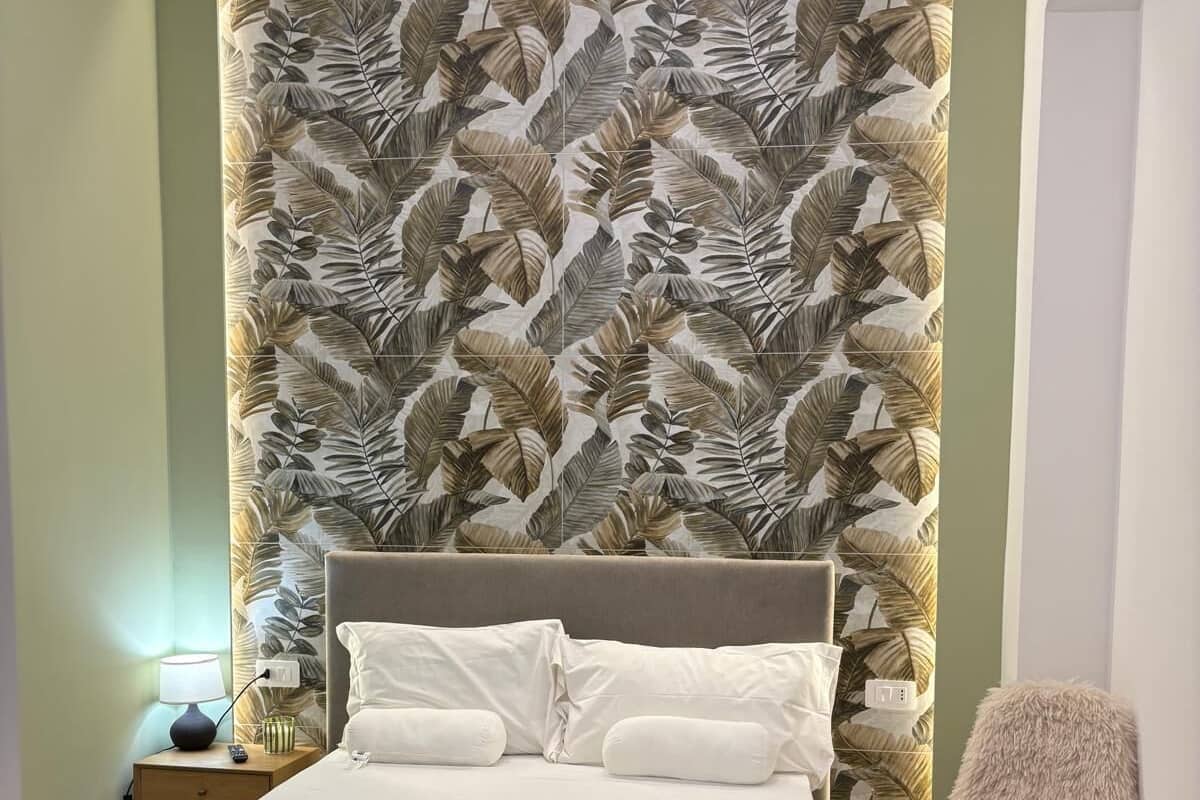 Ein Schlafzimmer mit einem Bett, einem Nachttisch, einer Lampe, einem Sessel und einer Wand mit Blättermotiv.