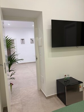Habitación de hotel con televisor de pantalla plana montado en la pared, una mesita auxiliar negra con adornos y una puerta que da a otra habitación.
