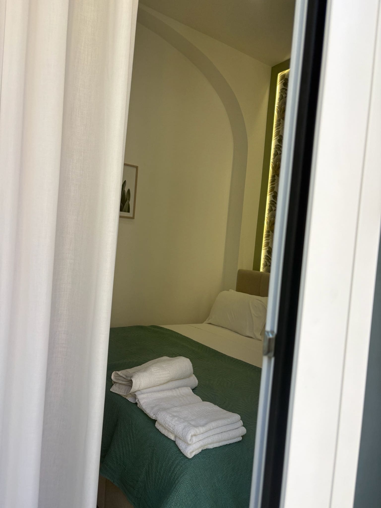 Una camera d'albergo con un letto, una coperta verde, cuscini bianchi e un asciugamano bianco piegato sul letto.